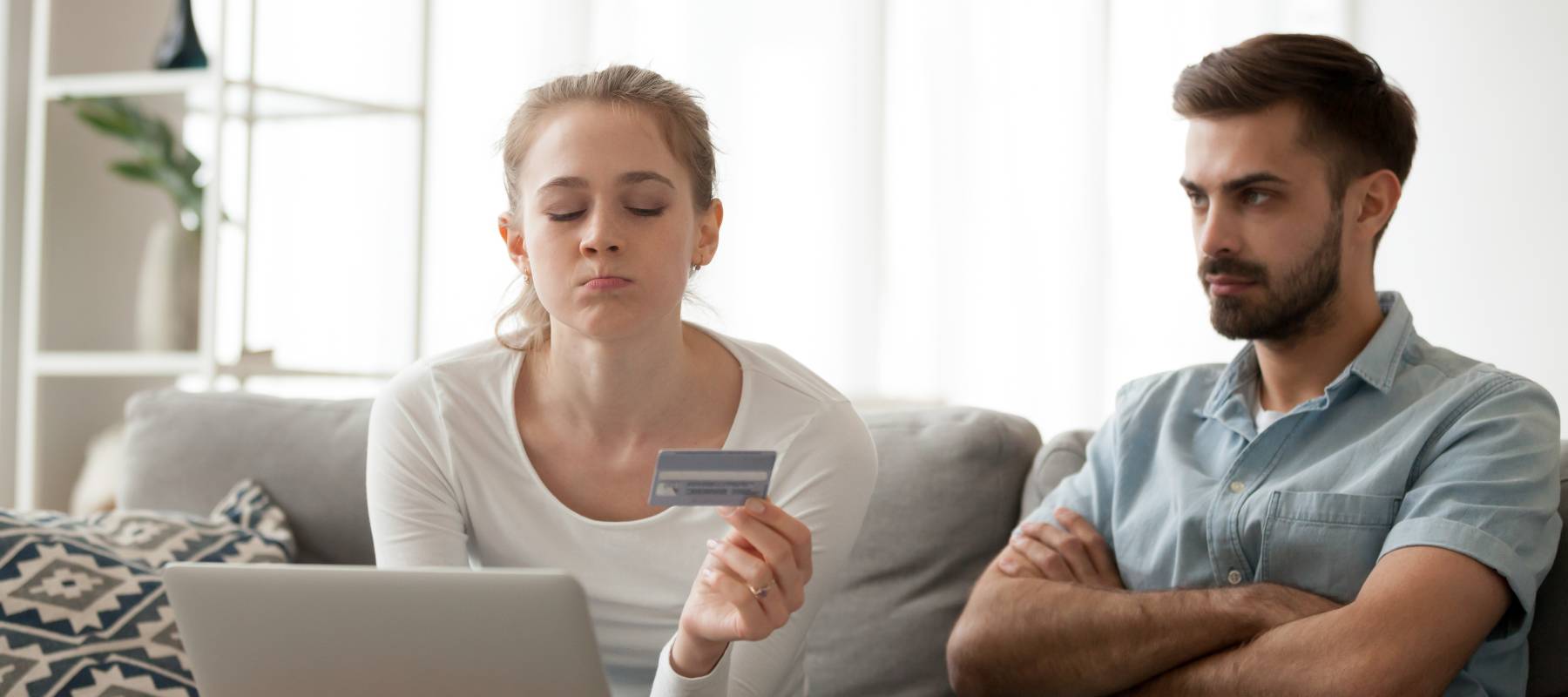 A woman and man sit on a grey couch in front of an open laptop. The woman purses her lips, eyes closed, as she holds a credit card at arm's length. The man stares into the distance furiously.