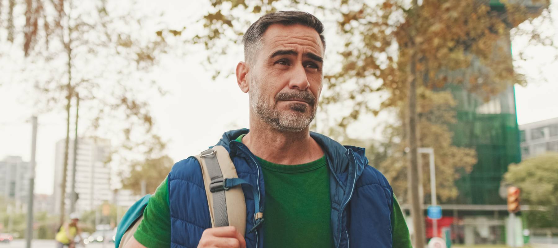 Middle aged man walking outside in casual clothes with one hand on a backpack strap.