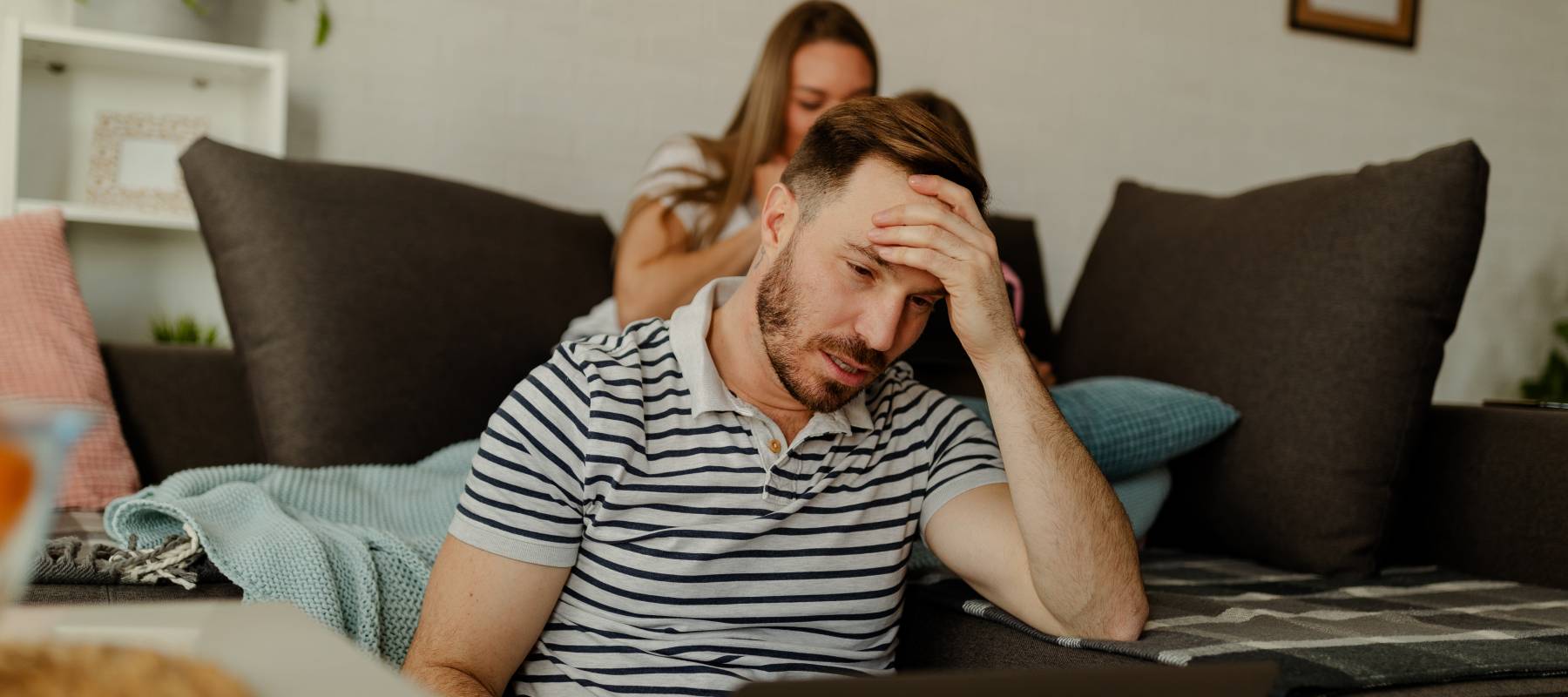 Frustrated young man sitting on the floor at home looking at computer in distress with wife in background on couch.