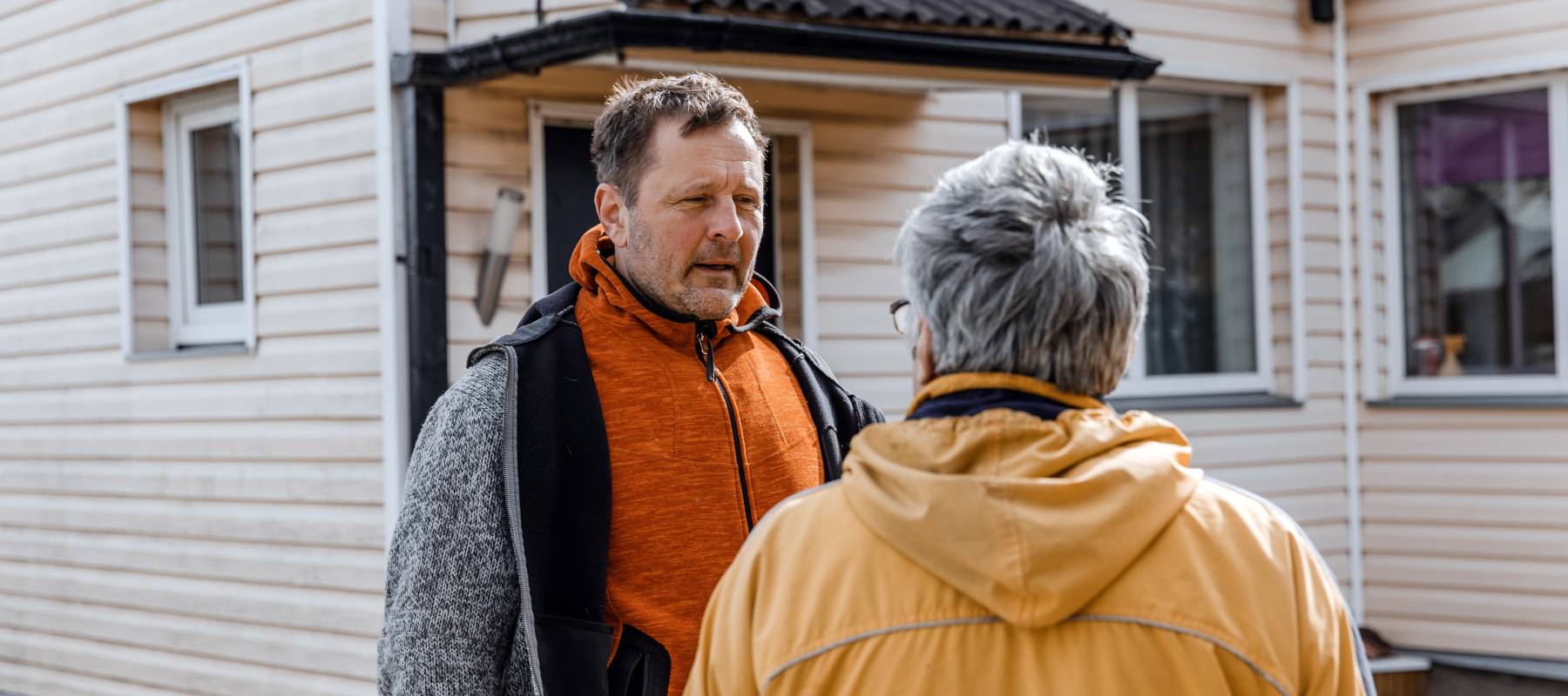 Man with white hair speaking to a younger gentleman outside a lake house.