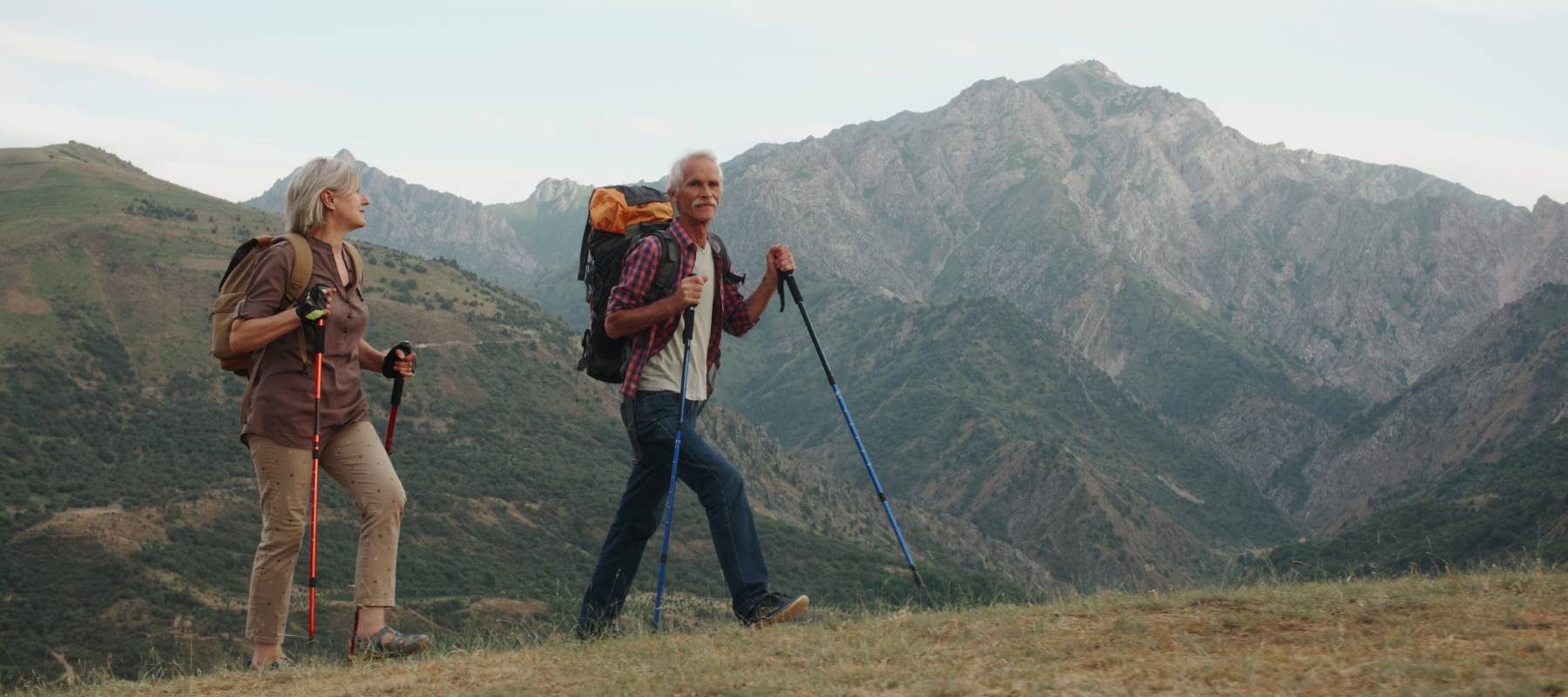 An older couple hike across a rise with mountains in the distance.