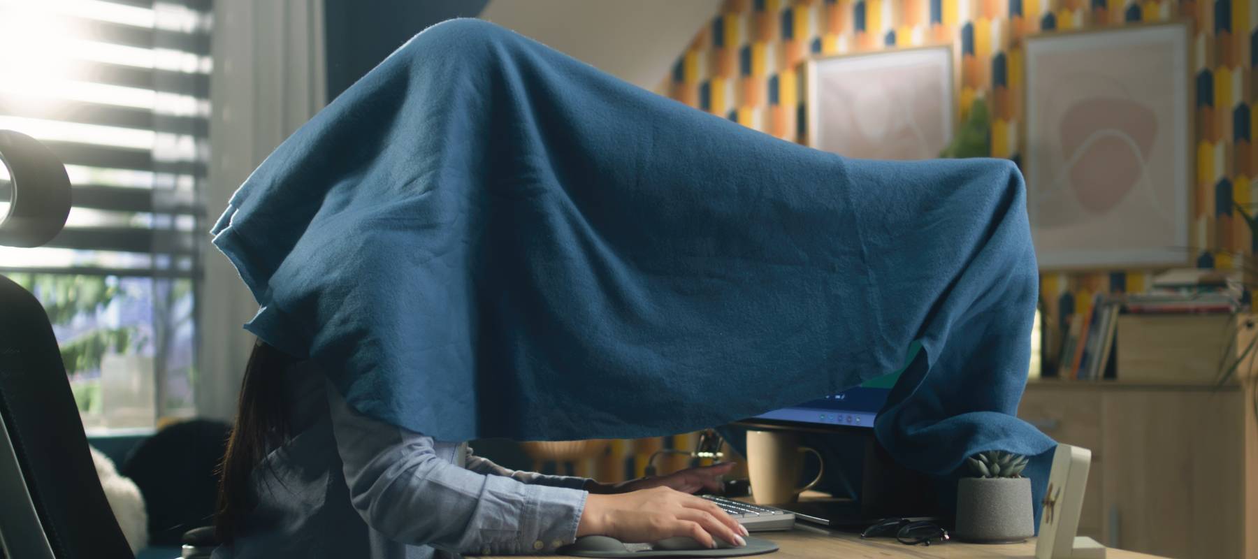 A person covered with a blanket over a computer and a desk.
