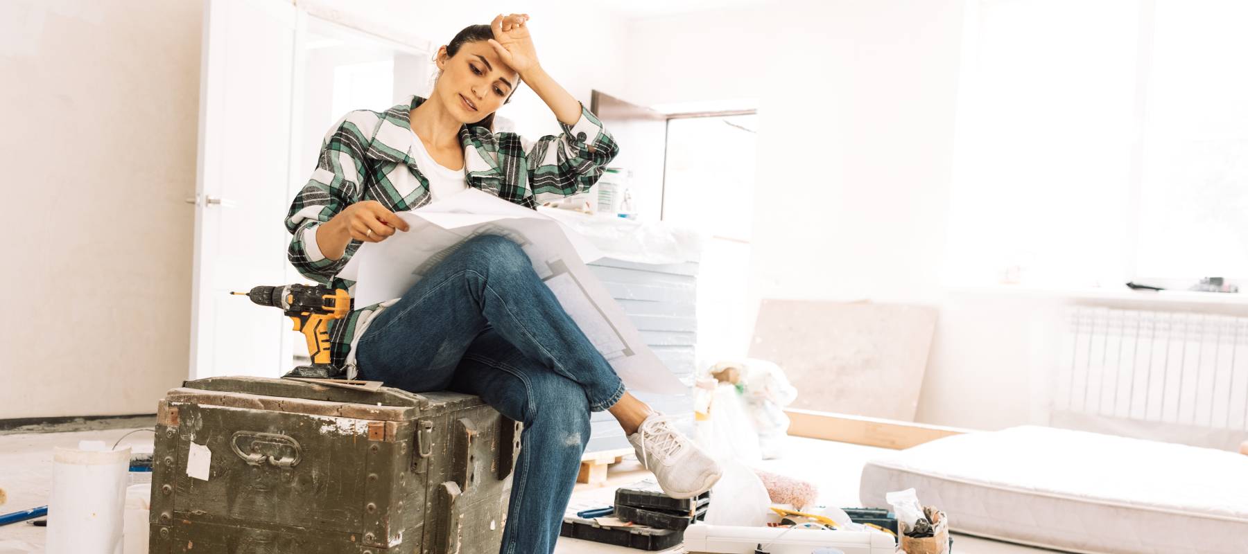 A woman in a flannel and jeans sits on a steamer's trunk with a cordless drill nearby and blueprints spread across one knee.