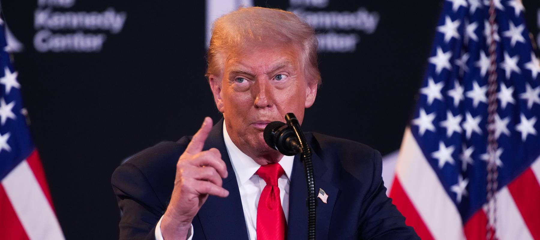 President Donald Trump holds up one finger while speaking at a podium with twin American flags in the background.