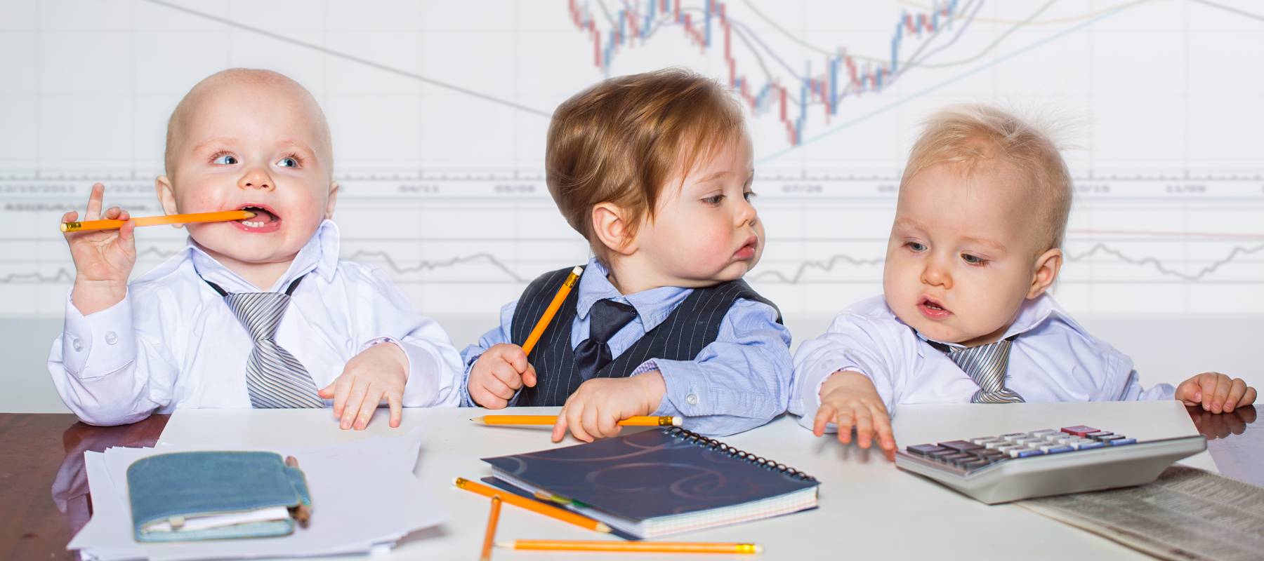 Three adorable babies sitting at a desk in business attire, in front of pencils, calculators and other office supplies.