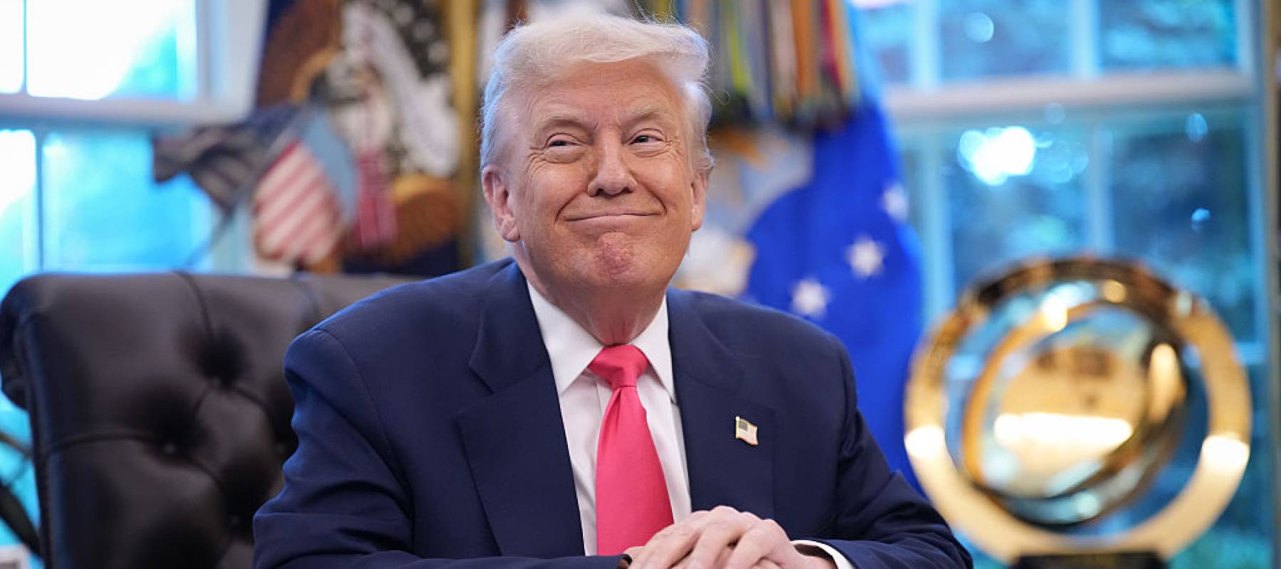 President Donald Trump smiles without showing his teeth at the Oval Office's iconic desk with a gold trophy in the background.