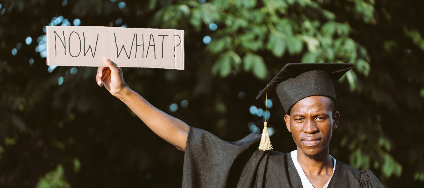 Young male graduate holding a cardboard sign that says, "Now what?"