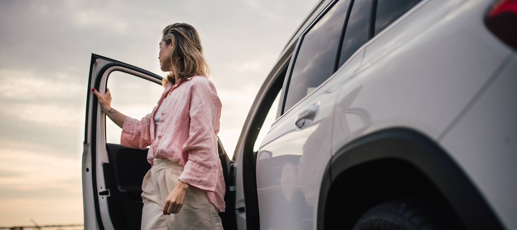 A woman in a pale pink blouse and beige linen pants steps out of a silver SUV with her face turned away from the viewer, looking to the horizon.