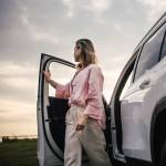 A woman in a pale pink blouse and beige linen pants steps out of a silver SUV with her face turned away from the viewer, looking to the horizon.