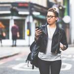 Girl walking down the street with her phone. A woman is walking alone in London. She is looking at her smart phone. Blurred on background there are  typical english houses and shops.