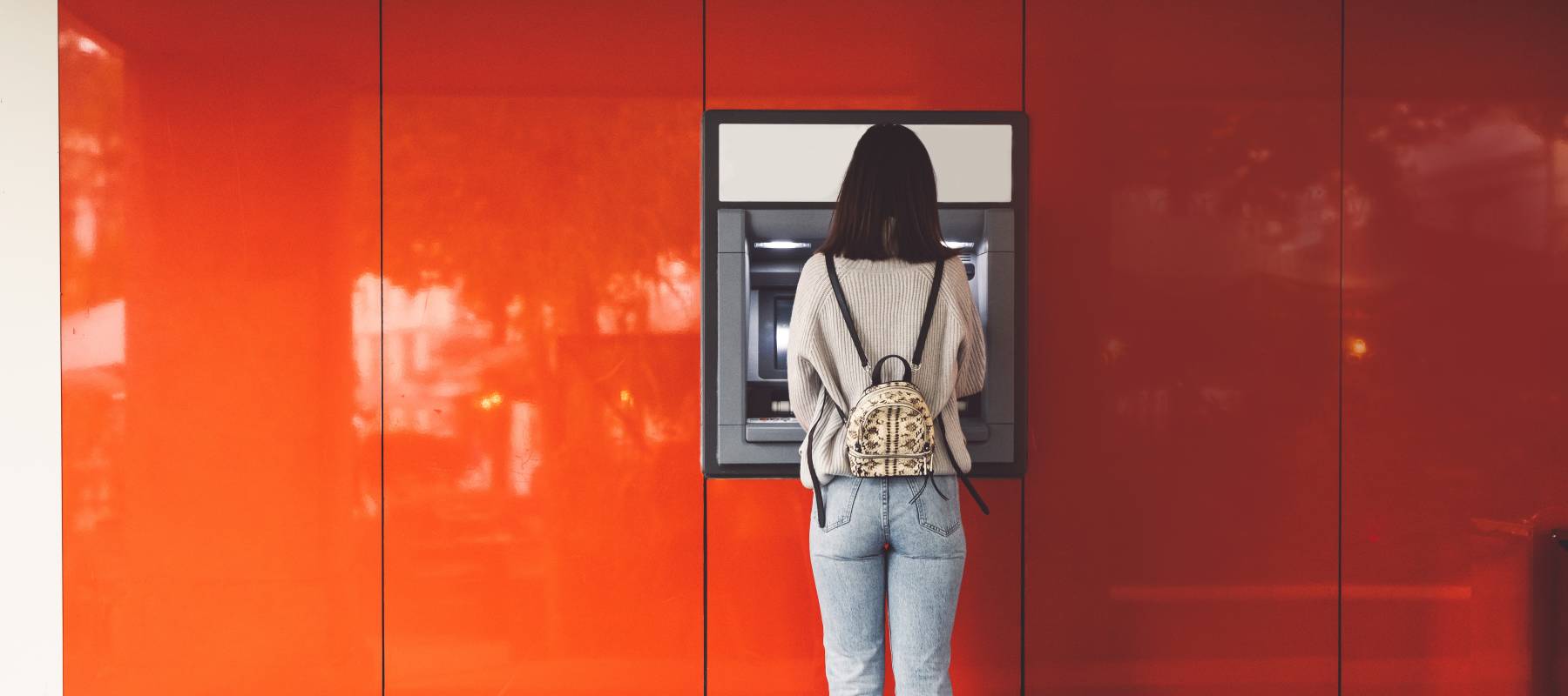 Back view of young woman standing in front of an ATM machine on a red wall
