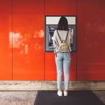 Back view of young woman standing in front of an ATM machine on a red wall