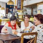 Three woman sit at a table in a coffee shop and look at a laptop with concern while sipping espresso and snacking.