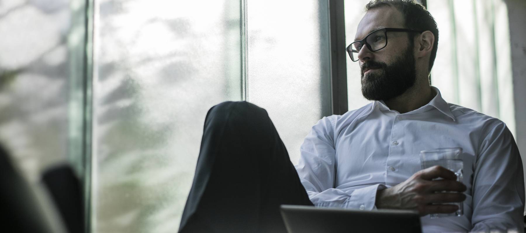 A man sits  with a glass of water looking out an office window.