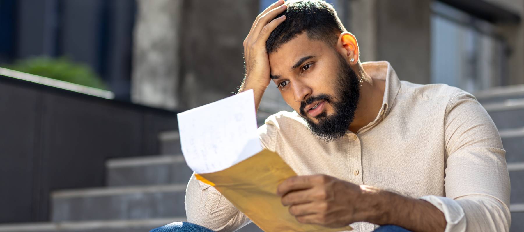 Worried man reading document seated on outdoor steps in a city setting.