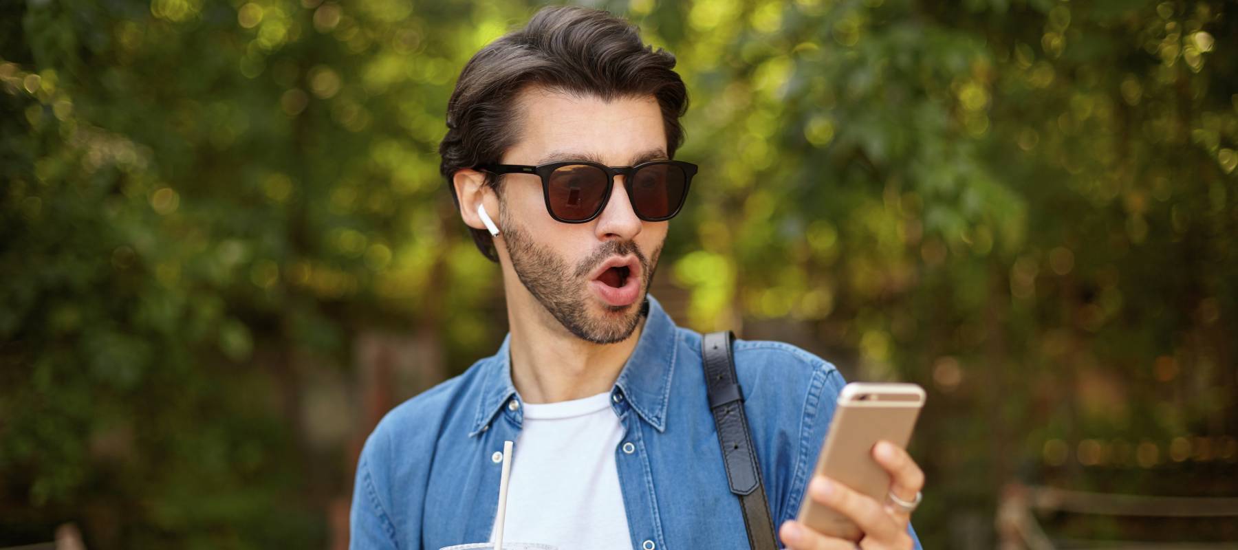 Surprised man in sunglasses standing over green park with mobile phone.