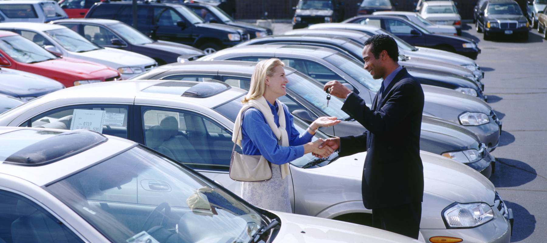 Woman buying car in large car sales parking lot while salesman hands her keys.