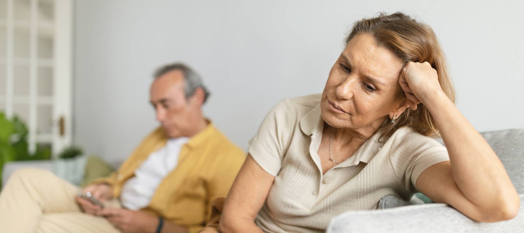 Woman looking disappointed and bothered on couch while man using smartphone in background.