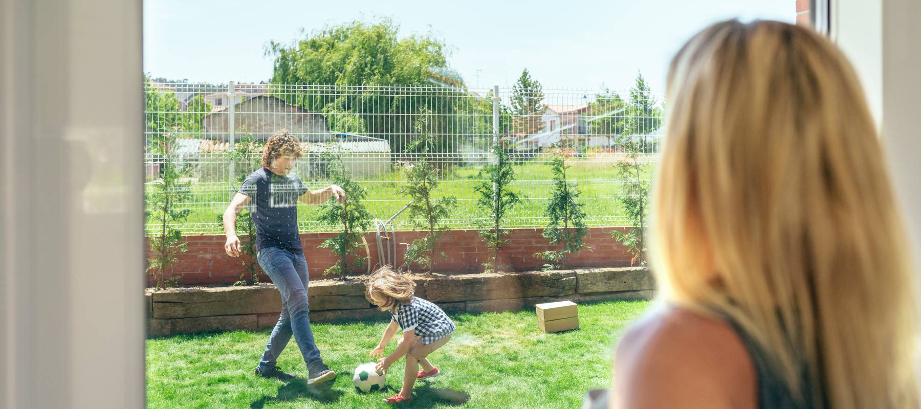 A blonde woman watching her teen son and young daughter playing soccer in the backyard through a window.