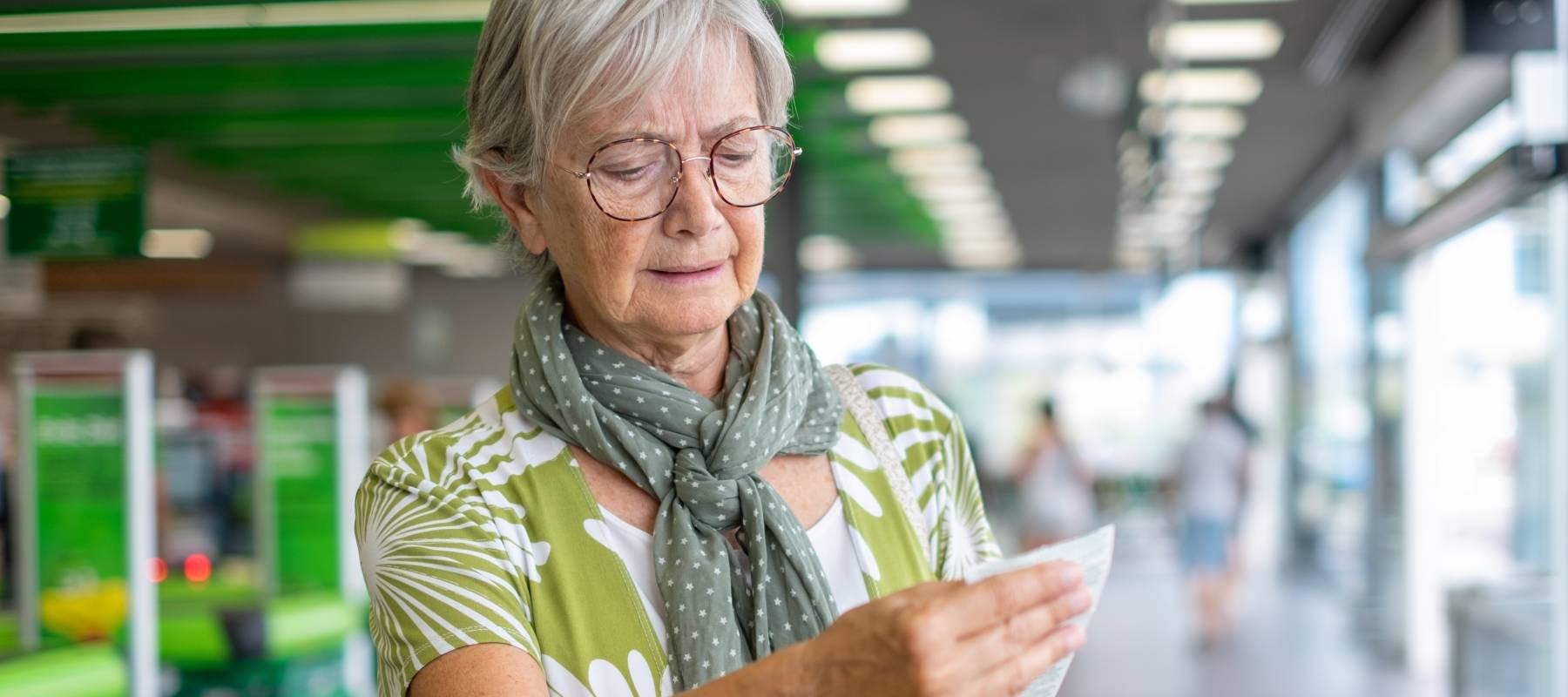 Senior woman in the supermarket checks her grocery receipt looking worried about rising costs.