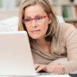 Woman wearing glasses lying on a sofa at home concentrating as she works on a laptop