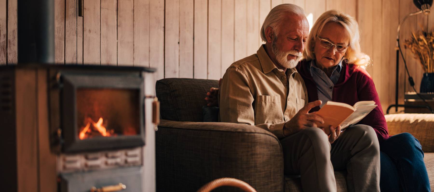 An elderly couple sitting by the fireplace, embracing, and enjoying a book together.