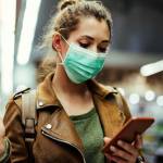 Young woman wearing protective mask on her face and reading shopping list on mobile phone in grocery store during virus pandemic.