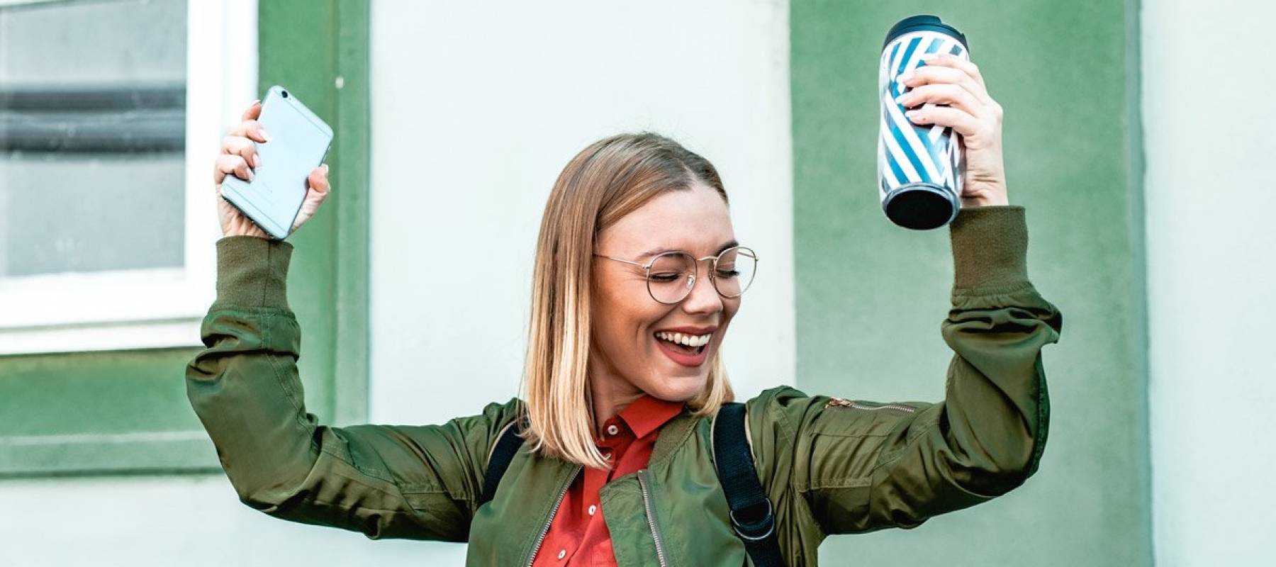 Happy young woman with short blonde hair and glasses holding her smartphone and coffee cup over her head