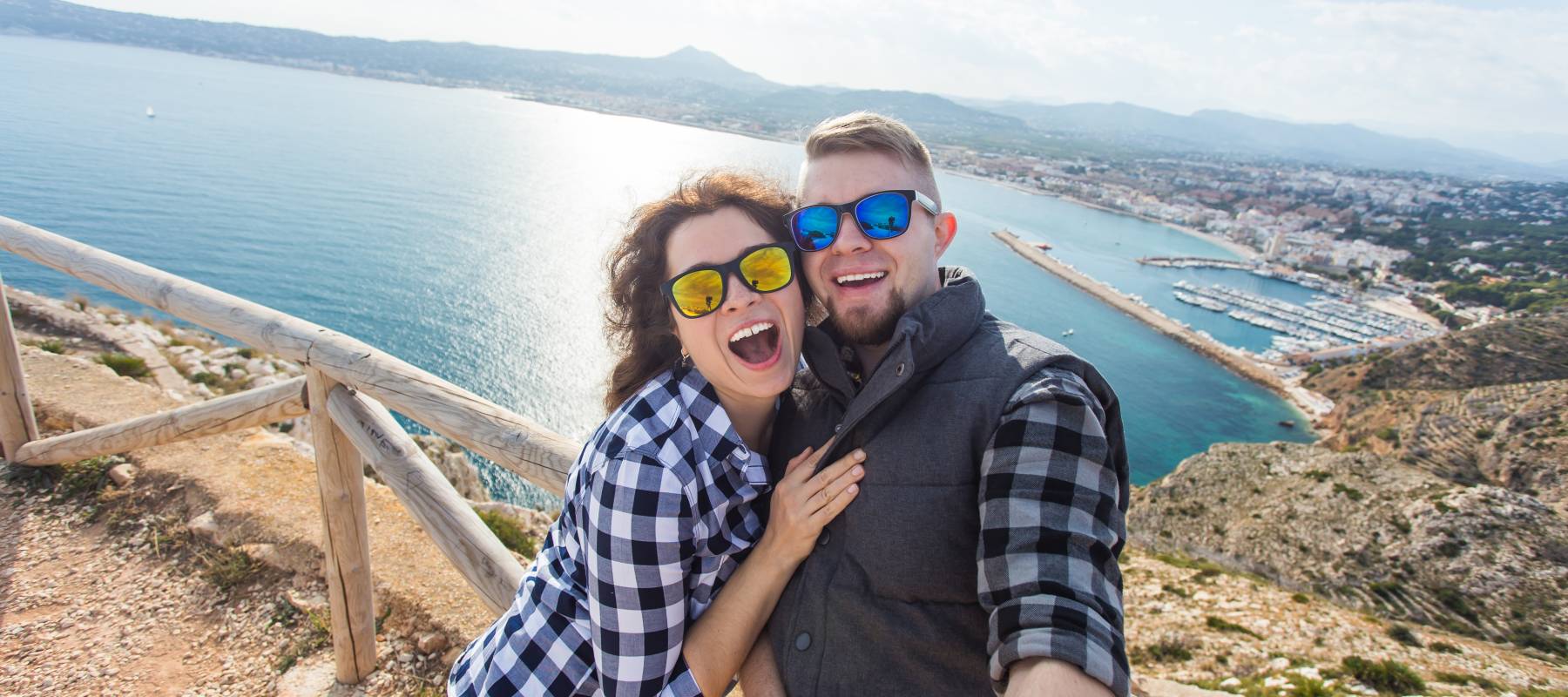 A couple wearing sunglasses and dressed in flannel take a selfie at a sandy shoreline with a breakwater and small port in the background --- mountains loom on a distant coast.