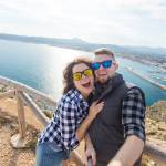 A couple wearing sunglasses and dressed in flannel take a selfie at a sandy shoreline with a breakwater and small port in the background --- mountains loom on a distant coast.
