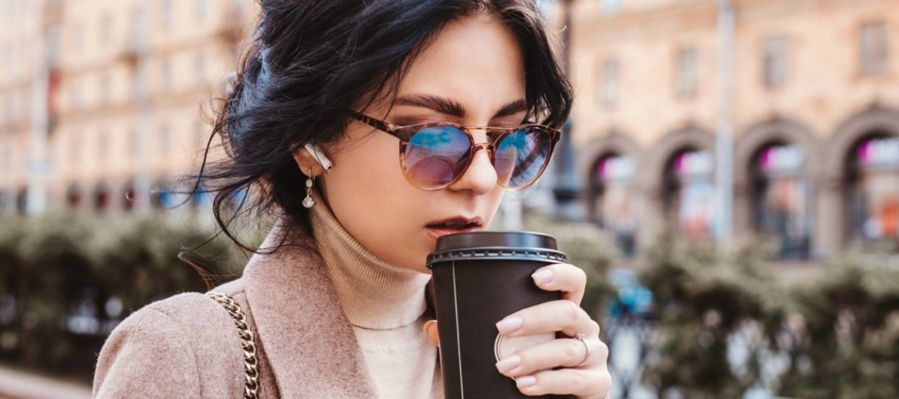 Woman with dark hair and sunglasses walking outdoors with takeout coffee