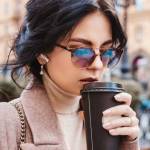 Woman with dark hair and sunglasses walking outdoors with takeout coffee