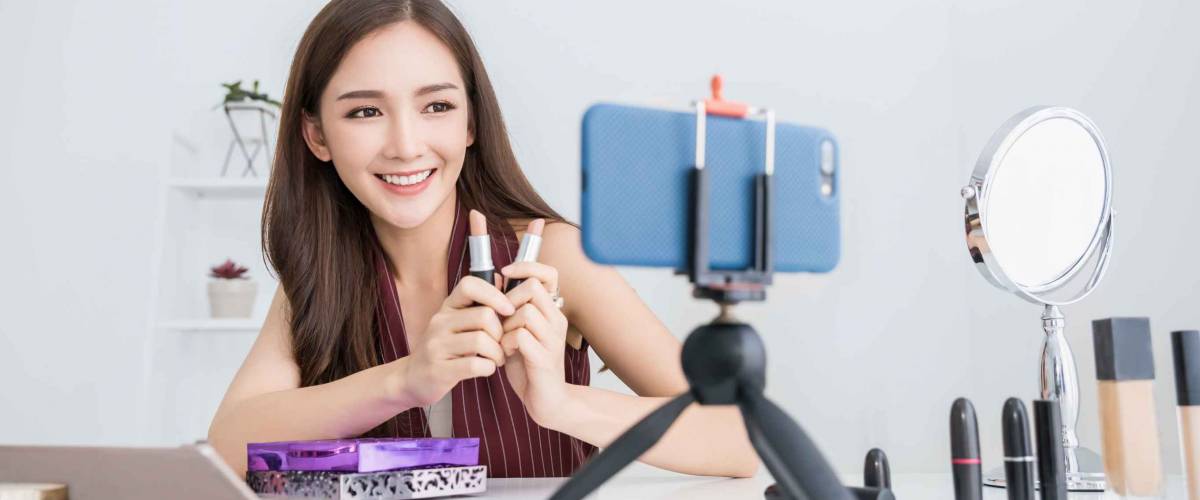 Young Asian-American woman making video about lipsticks on smartphone for posting to YouTube