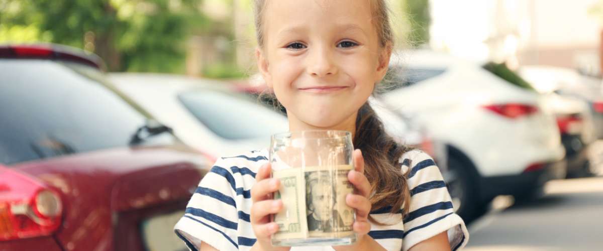 How To Teach Your Kids About Saving Money - cute girl holding bank with money in hands and sitting in shopping cart outdoors