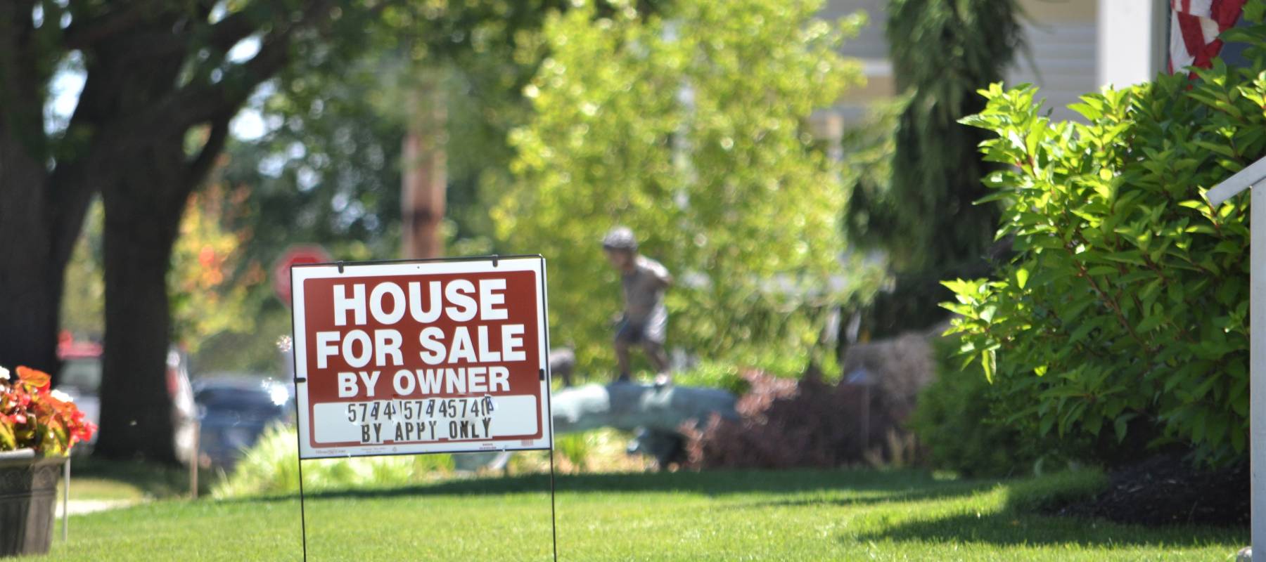 A "For Sale" sign sits on a law in a suburban neighbourhood, background racked slightly our of focus, on a crisp summer's day.