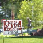 A "For Sale" sign sits on a law in a suburban neighbourhood, background racked slightly our of focus, on a crisp summer's day.