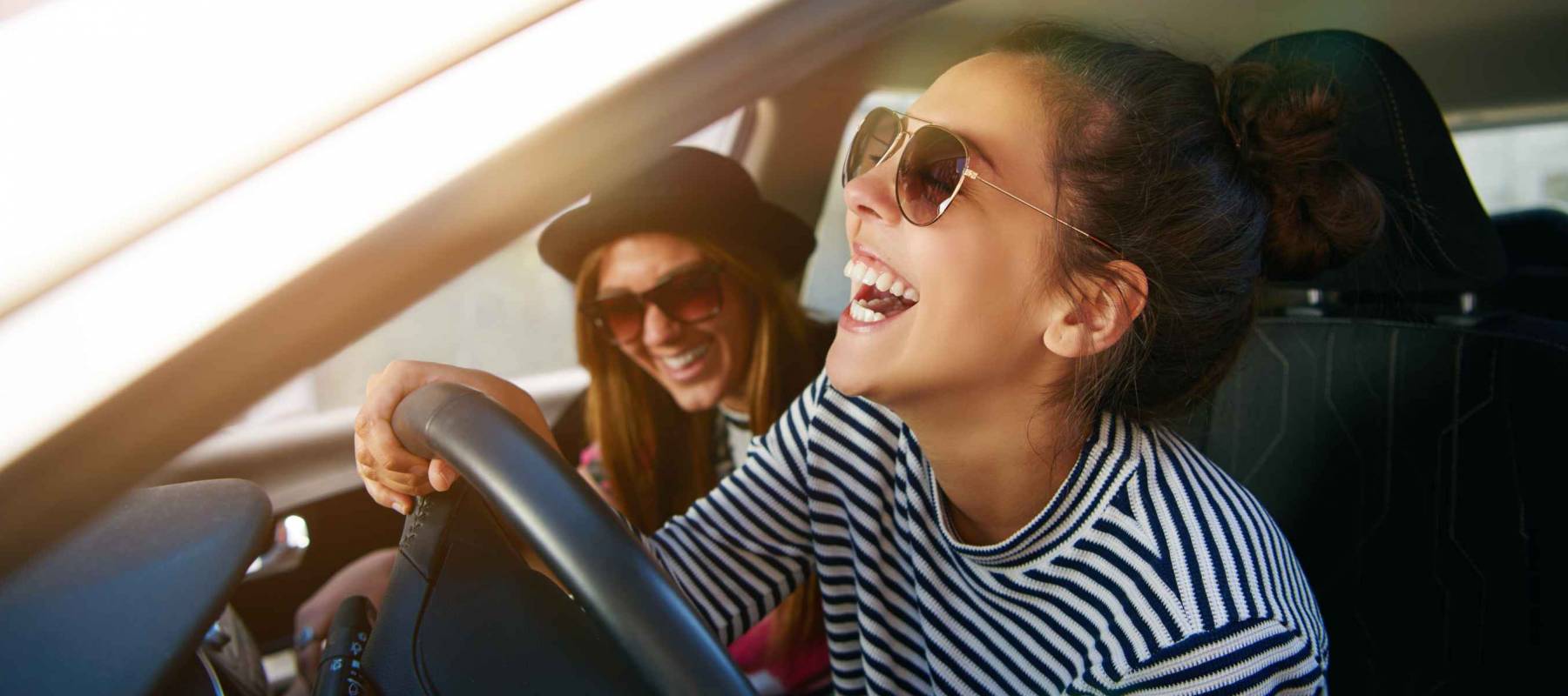 Laughing young woman wearing sunglasses driving a car with her girl friend , close up profile view through the open window