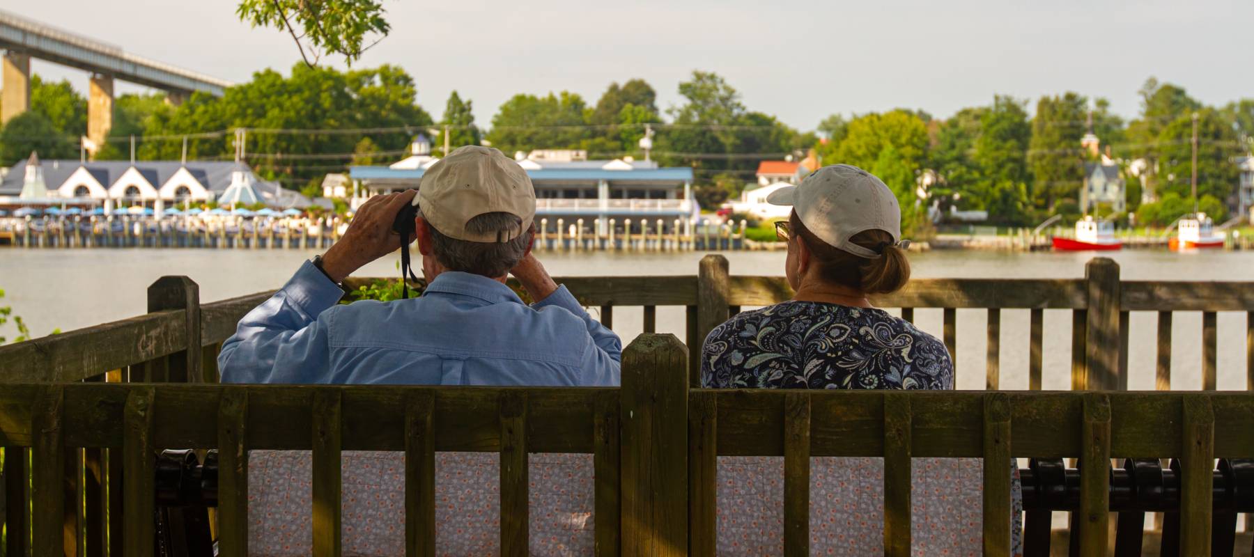 Older couple wearing baseball hats sitting on a wooden outdoor bench in a garden deck by the Chesapeake and Delaware Canal.