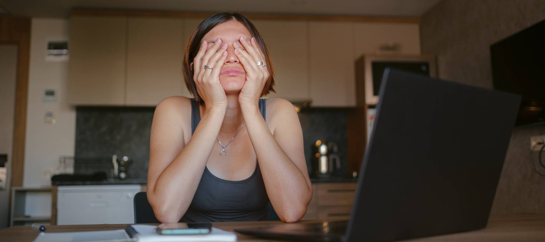 Woman sitting at a kitchen table with a laptop with her head in her hands.