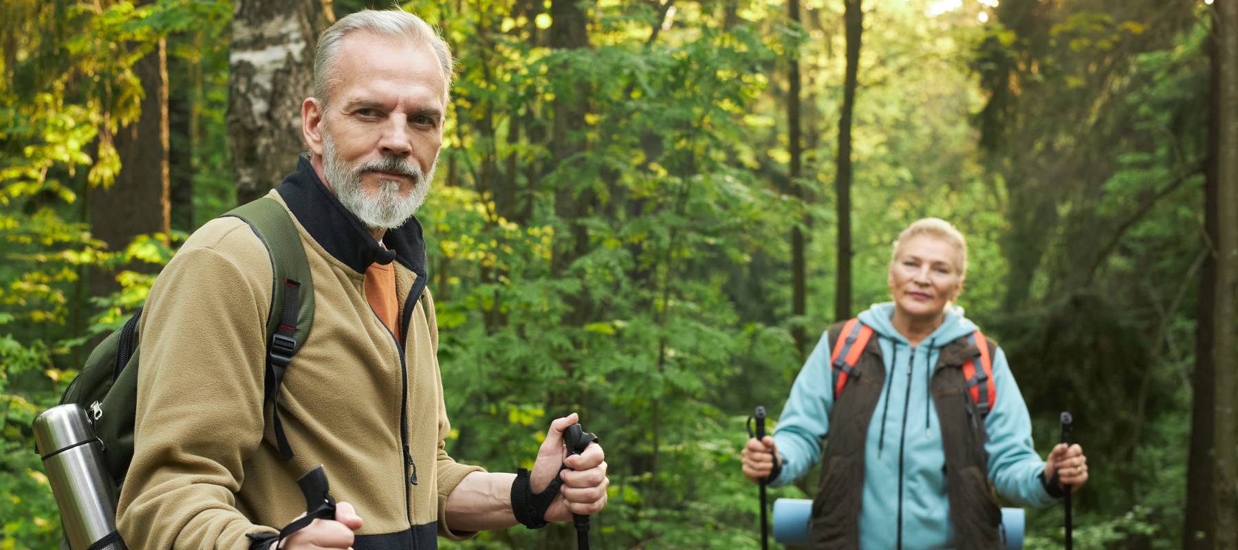 Senior Caucasian man and woman hiking with trekking poles in forest.