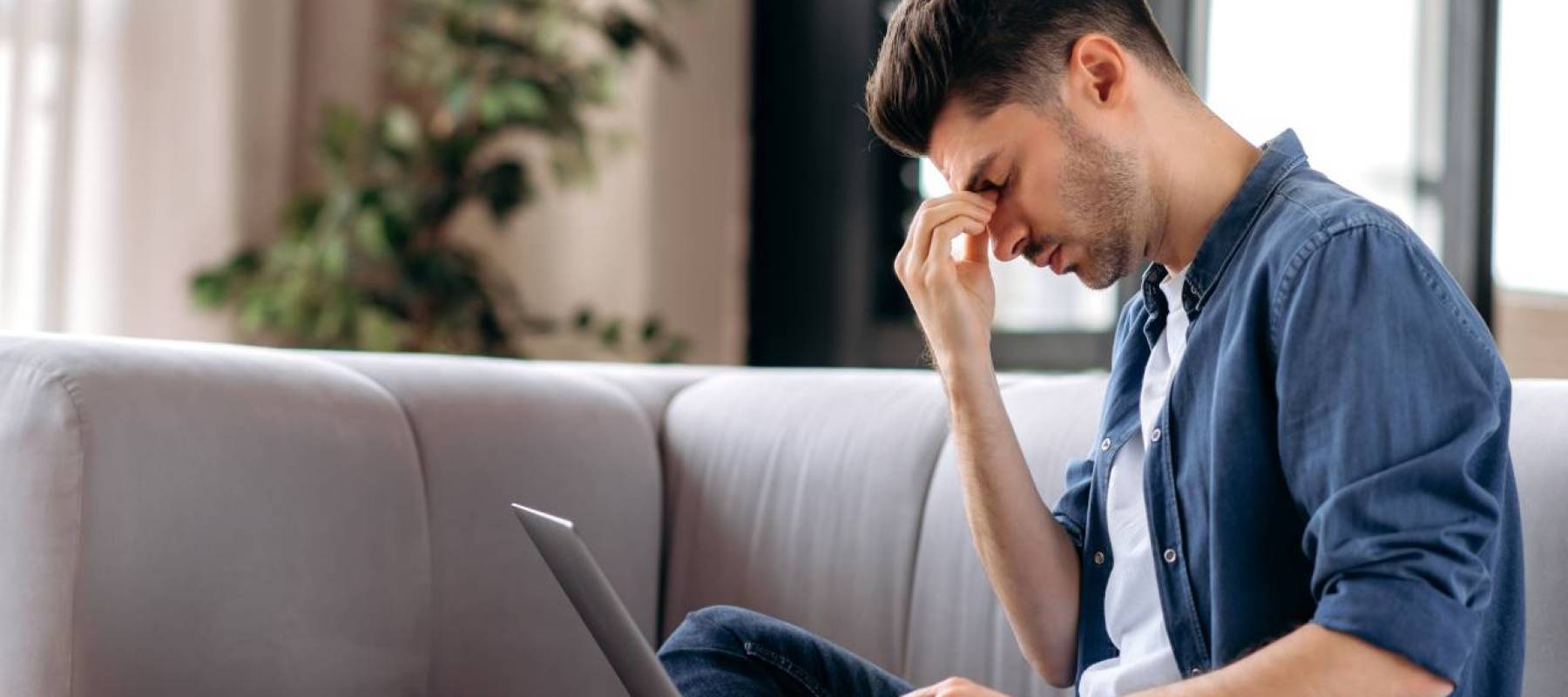 young man sits on couch with computer looking upset