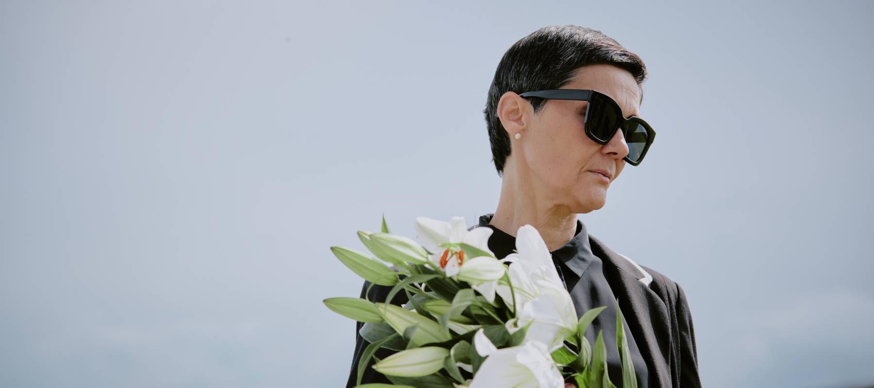 middle aged Caucasian woman wearing sunglasses holding lilies at a funeral