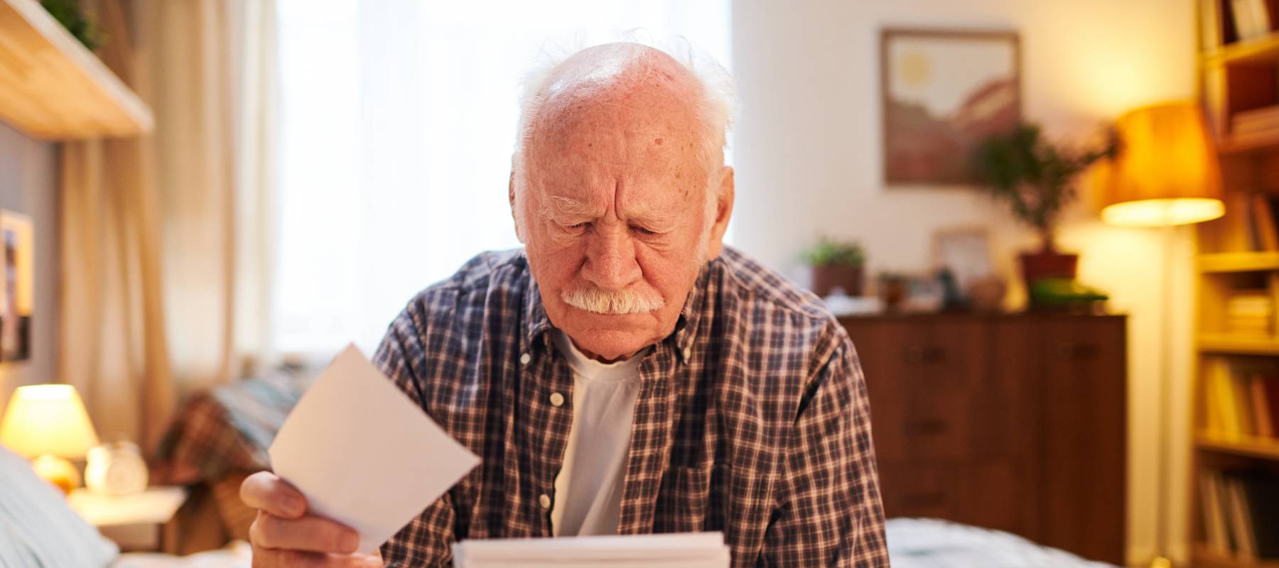 Old man sitting on the bed in bedroom and looking at photos in his hands