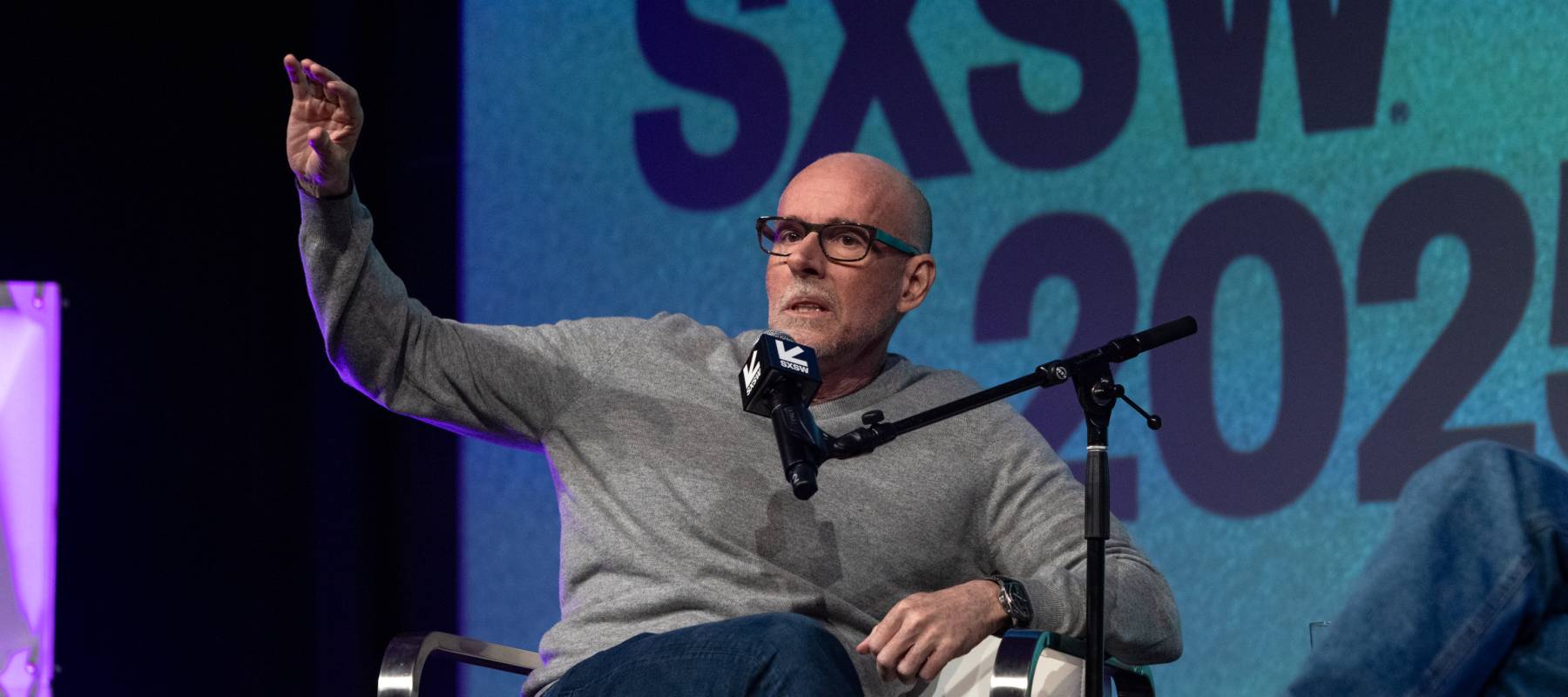 Prof. G, wearing a grey sweater and thick black framed glasses, raises a hand and looks askew at the camera.