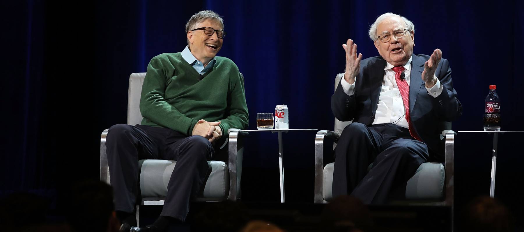 Warren Buffett raises his hands to the audience while Bill Gates lets out a short laugh.