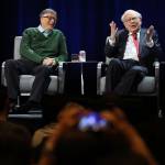 Warren Buffett raises his hands to the audience while Bill Gates lets out a short laugh.