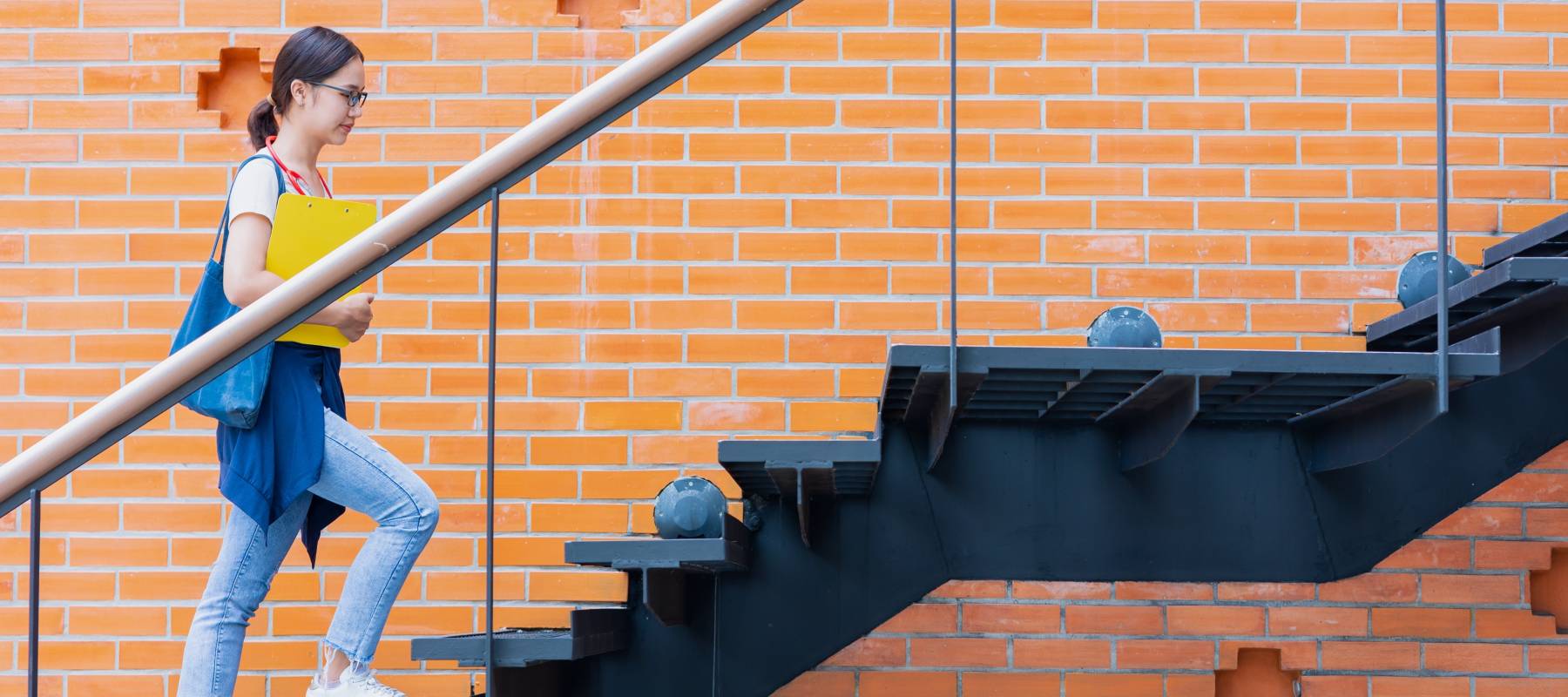 young asian woman walks alone up stairs wearing backback on background of bright orange brick wall