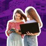 Two college students are standing near the university, looking at a computer, making notes in a notebook.