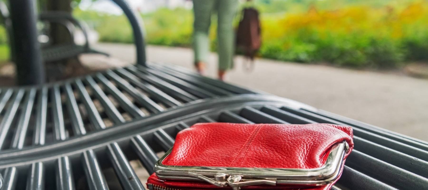 Someone walking away from a park bench, having left a red pocketbook behind.