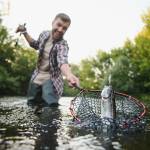 Man with fishing rod, catching a catfish in a net, close up, standing in a river outdoors.